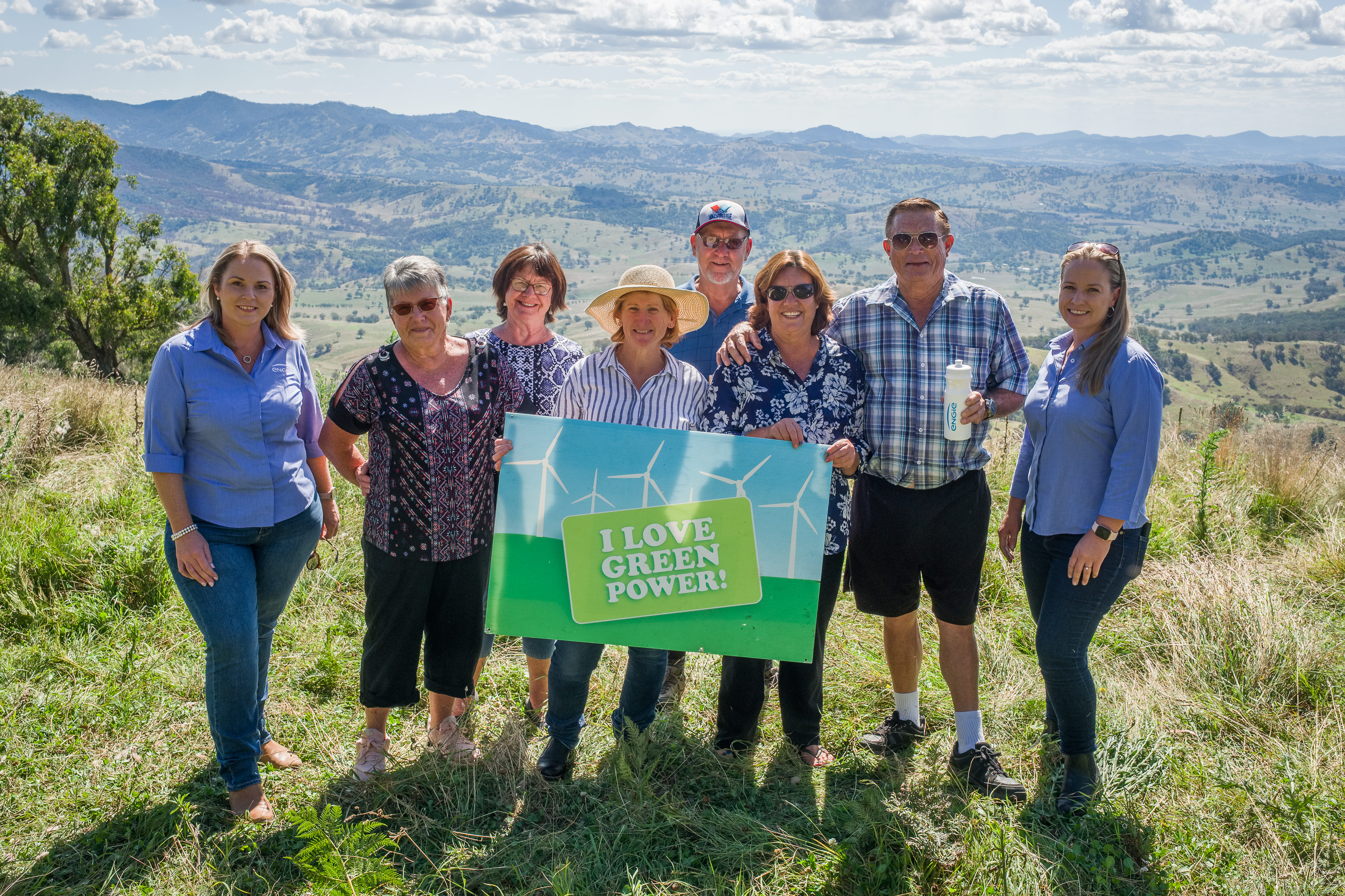 Group of happy, smiling community members holding an 'I love green power' sign. View of mountains in the background