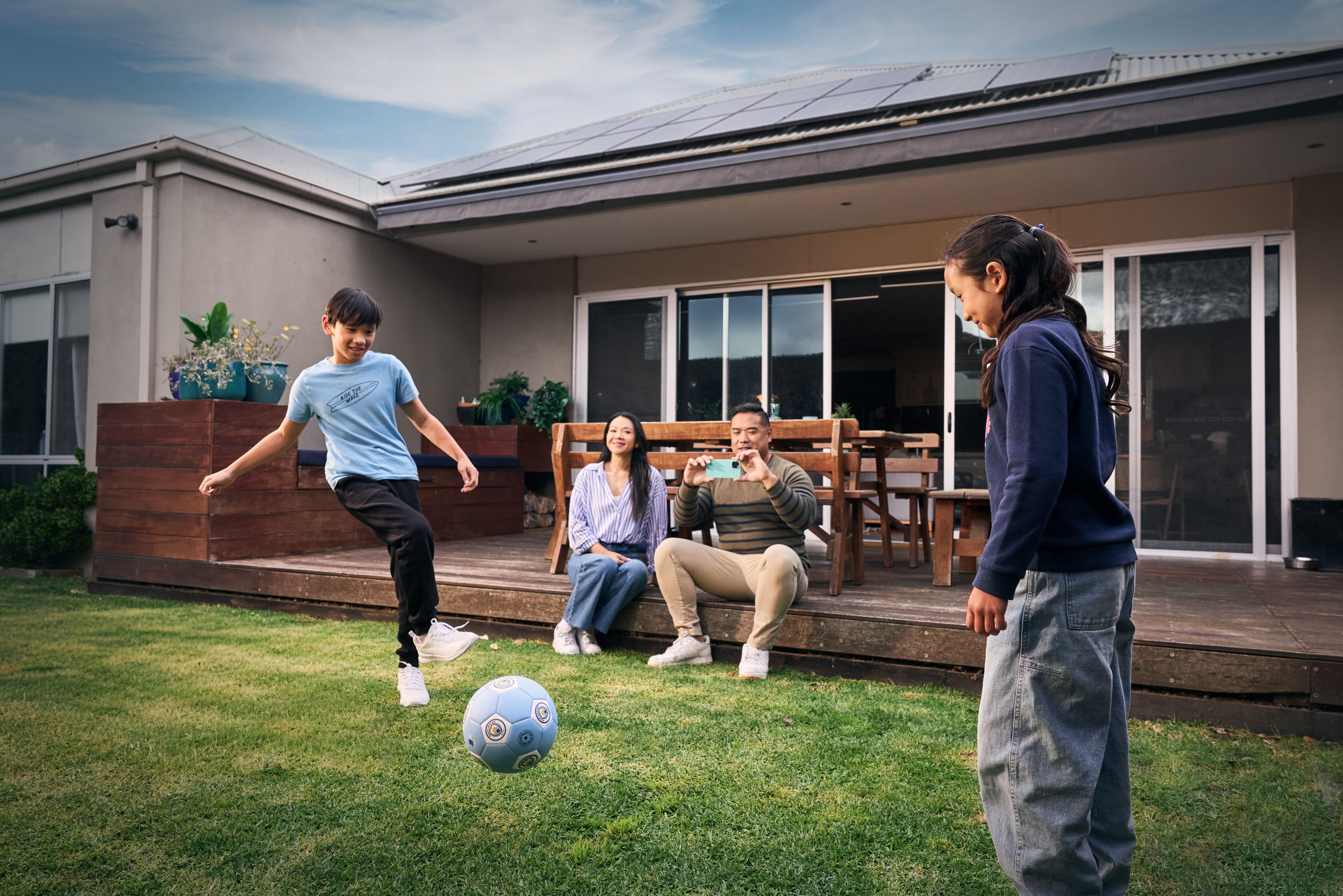 Family in their back yard. Two children are playing soccer on the grass. Mum and dad are sitting on the deck and dad is taking photos with his mobile phone.