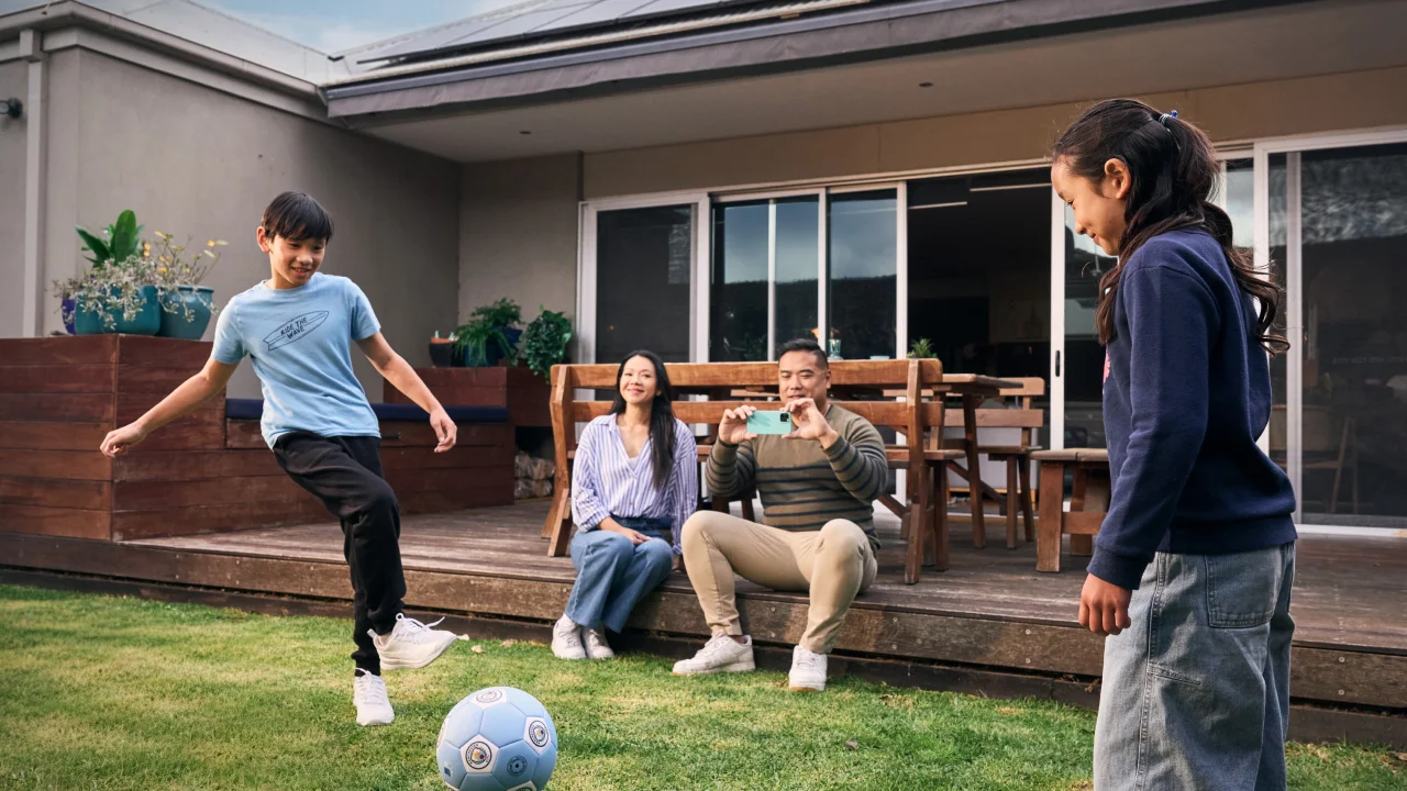 Two kids playing soccer in the backyard while their parents watch and take photos, enjoying family time at home.