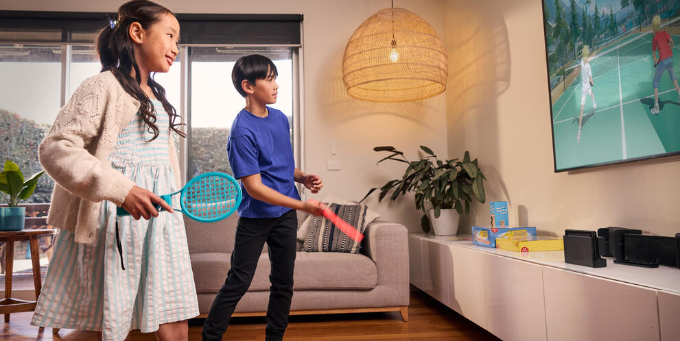 Two children are playing a motion-controlled tennis video game in a living room using handheld controllers