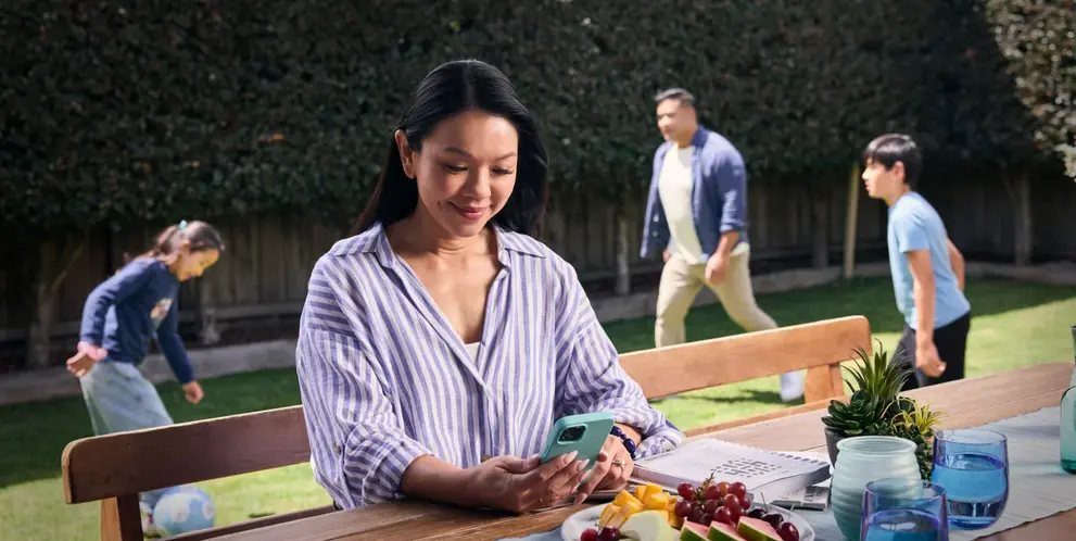 A person sitting at an outdoor table using a smartphone while children play in the backyard.