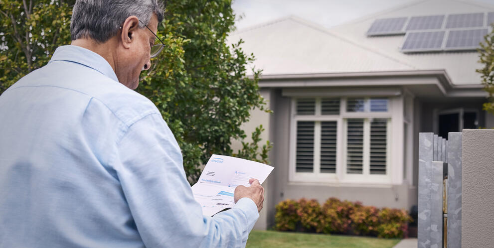 A person stands outside a house with rooftop solar panels, holding and looking at a utility bill near the front gate.