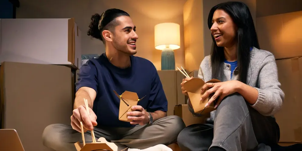 Two people sitting among moving boxes eating takeaway.