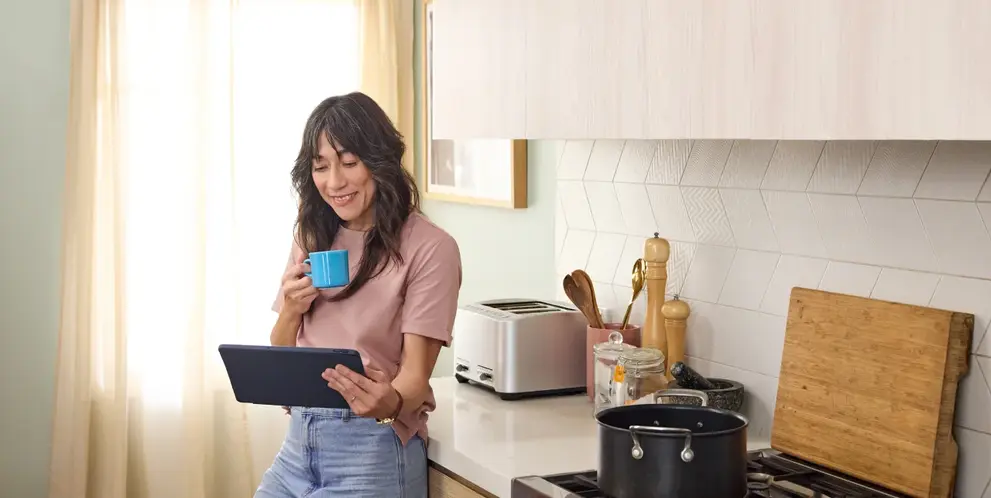 A person standing in a kitchen holding a tablet and drinking from a mug, with cooking items on the counter and a pot on the stove.