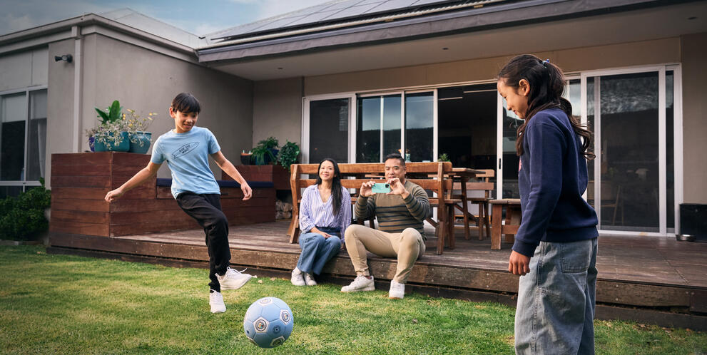 Two children playing soccer on a grassy backyard while two people sit on a deck. The house in the background has solar panels on the roof.