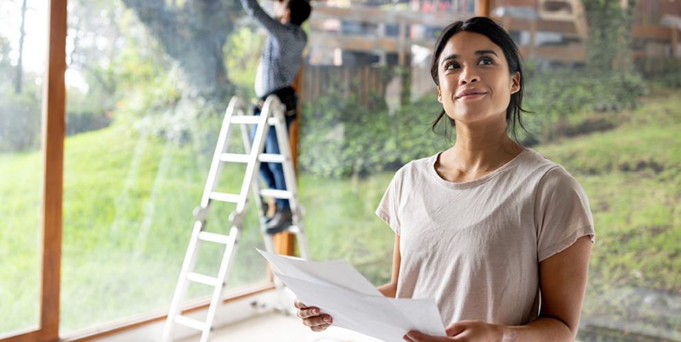Person holding documents inside a home while another person works on a large window using a ladder.