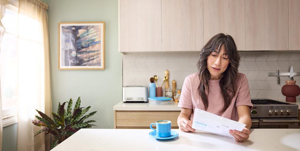 Person sits at a kitchen counter reading a bill with a cup of coffee nearby
