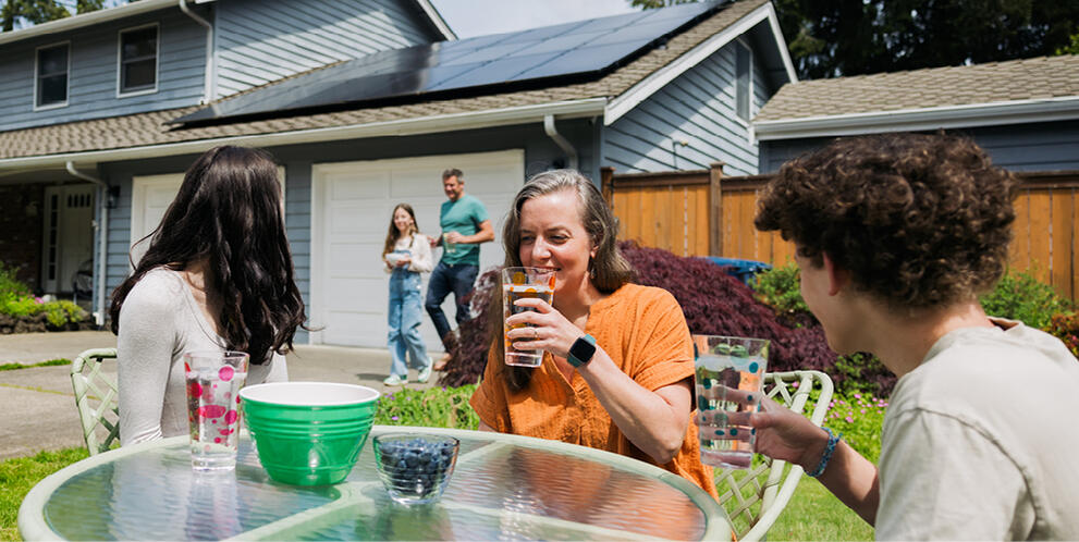 Group of people sitting at an outdoor table with drinks and snacks in a backyard, with a house featuring rooftop solar panels in the background