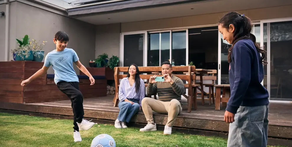 Two kids playing soccer in the backyard while their parents watch and take photos, enjoying family time at home.