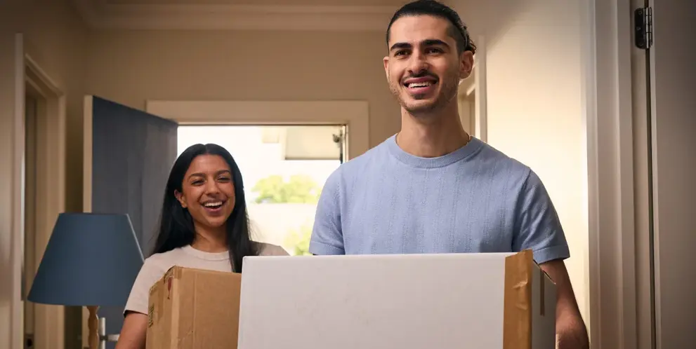 A smiling couple carrying moving boxes into a new home. ENGIE's moving home guarantee means their new home is already connected to energy.
