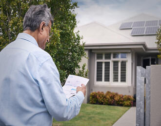 A person stands outside a house with rooftop solar panels, holding and looking at a utility bill near the front gate.