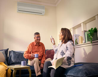 Two people sit on a couch; one holds a remote, the other a mug. An air conditioner is mounted above, with books and a plant on a shelf behind