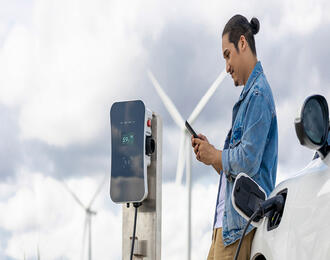 A person stands beside a white electric vehicle while it charges at an outdoor charging station, using a smartphone. Wind turbines spin in the background under a cloudy sky.