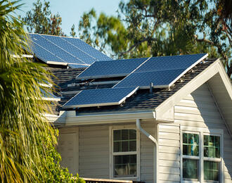 Solar panels installed on the roof of a light‑coloured house surrounded by trees and greenery.