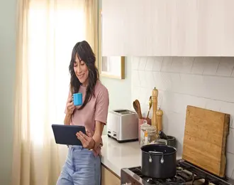 A person standing in a kitchen holding a tablet and drinking from a mug, with cooking items on the counter and a pot on the stove.