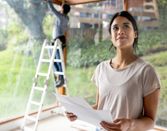 Person holding documents inside a home while another person works on a large window using a ladder.