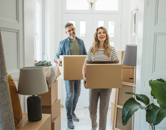 Two people carrying cardboard boxes inside a bright room filled with packed items, including a lamp, cushions, and a large green plant