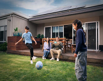 Two children playing soccer on a grassy backyard while two people sit on a deck. The house in the background has solar panels on the roof.
