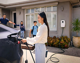 A woman plugging an electric vehicle charger into a dark car outside a house while a family walks out of the front door in the background.