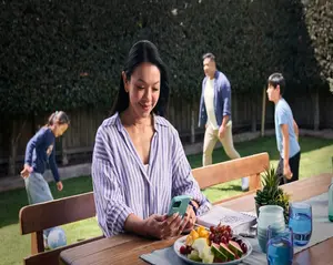 A person sitting at an outdoor table using a smartphone while children play in the backyard.