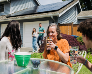 Group of people sitting at an outdoor table with drinks and snacks in a backyard, with a house featuring rooftop solar panels in the background
