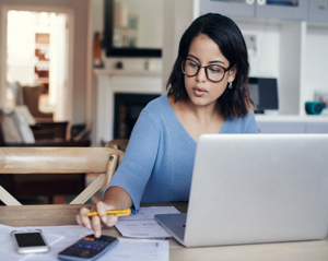 Person sitting at a wooden table working on a laptop with papers, a smartphone, and a calculator spread out, inside a home office or living space